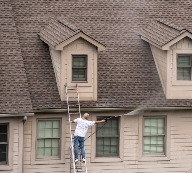 Painter on ladder using pressure wash spray to clean woodwork before painting townhome, siding water damage, mold on house exterior, algae growth on siding, home exterior maintenance, water damage signs, house mold and mildew, dirty vinyl siding, exterior home repair, weather damage on siding, house in need of cleaning